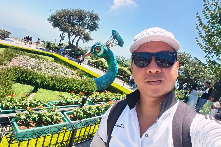 Viet Nguyen taking a selfie beside colorful peacock flower displays at the flower garden in Ba Na Hills during Ba Na Hills Golden Bridge tour.